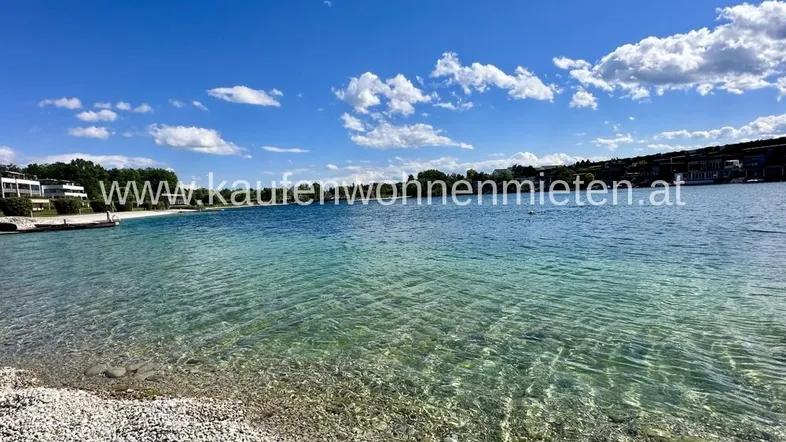 Klarer See mit Kieselstrand und grüner Uferlandschaft unter blauem Himmel.