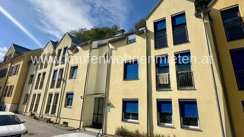 Modernes Mehrfamilienhaus mit gelber Fassade, kleinen Balkonen und blauen Rollläden unter blauem Himmel.
