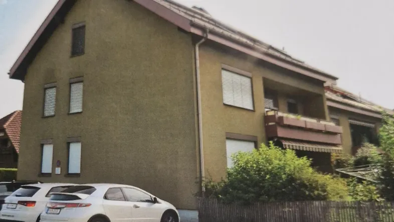 Gebäudeansicht mit Satteldach, Balkon und geparkten Autos auf der Straße.