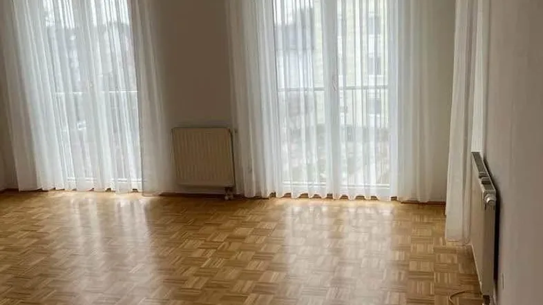 Helles Wohnzimmer mit Parkettboden und zwei großen Fenstern, die viel Tageslicht hereinlassen.