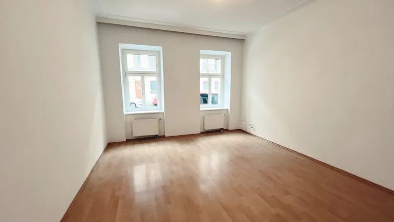 Helles, leeres Zimmer mit Parkettboden und zwei Fenstern, bereit zur individuellen Gestaltung.