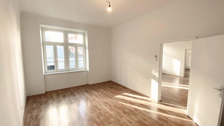 Helles Zimmer mit Holzfußboden und großem Fenster, das viel Tageslicht hereinlässt.