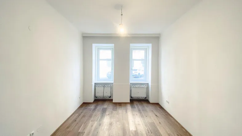 Helles, leeres Zimmer mit Holzboden und zwei Fenstern, bereit zur individuellen Gestaltung.