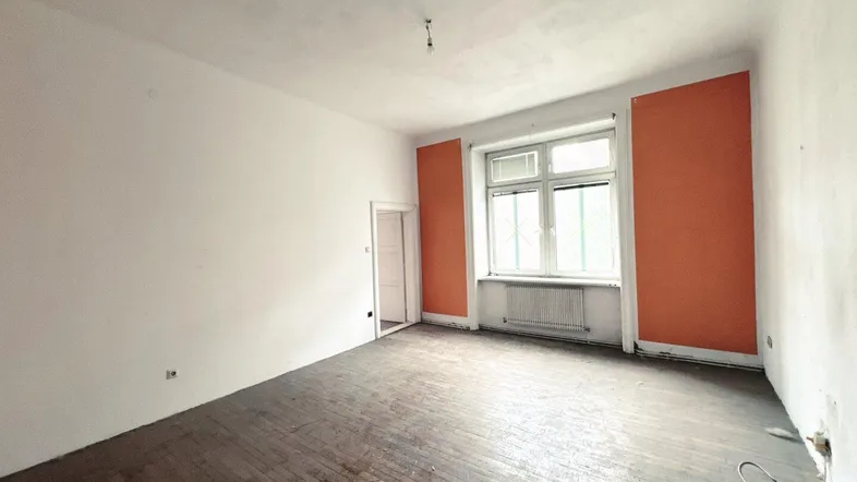 Geräumiges Zimmer mit abgenutztem Holzboden und orangefarbenen Akzentwänden, bereit zur Renovierung.