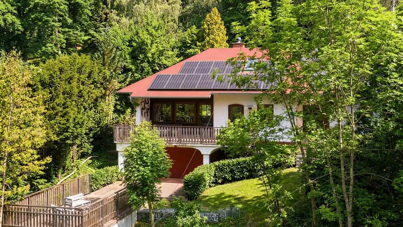 Freistehendes Haus mit rotem Dach und Solarpaneelen, umgeben von üppiger grüner Vegetation.