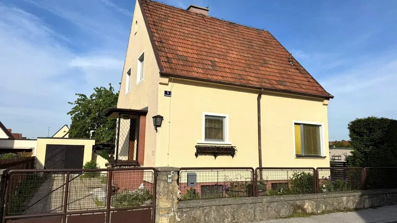 Gelbes Einfamilienhaus mit rotem Ziegeldach und Vorgarten, umgeben von einem Zaun.