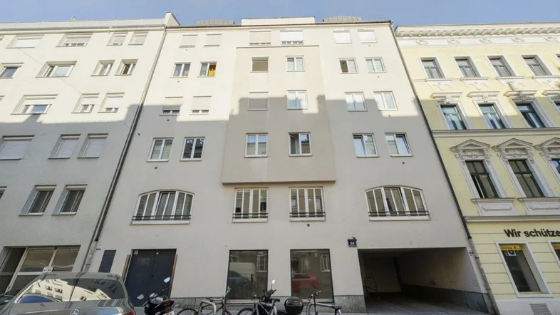 Ansicht der Gebäudefassade mit mehreren Stockwerken und geparkten Fahrzeugen davor in urbaner Umgebung.