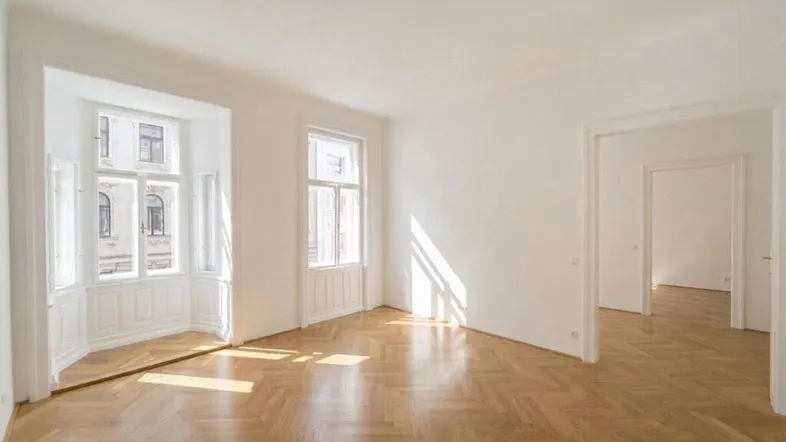 Helles, leeres Zimmer mit Fischgrätparkett und großen Fenstern, die viel Tageslicht hereinlassen.