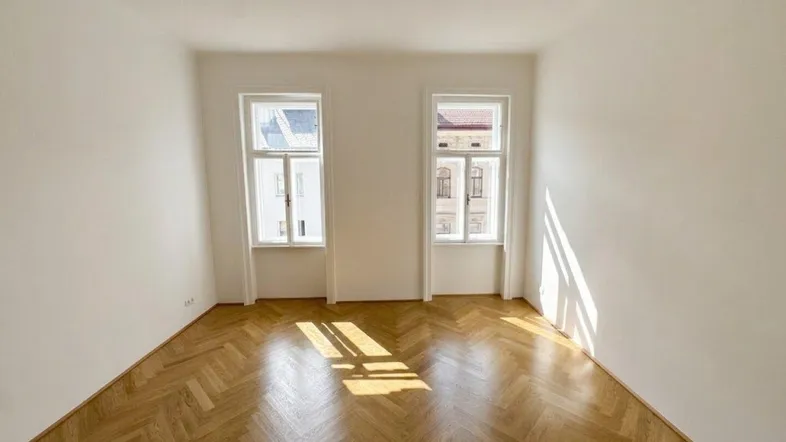 Helles, leeres Zimmer mit Fischgrätparkett und zwei Fenstern, das viel Tageslicht hereinlässt.