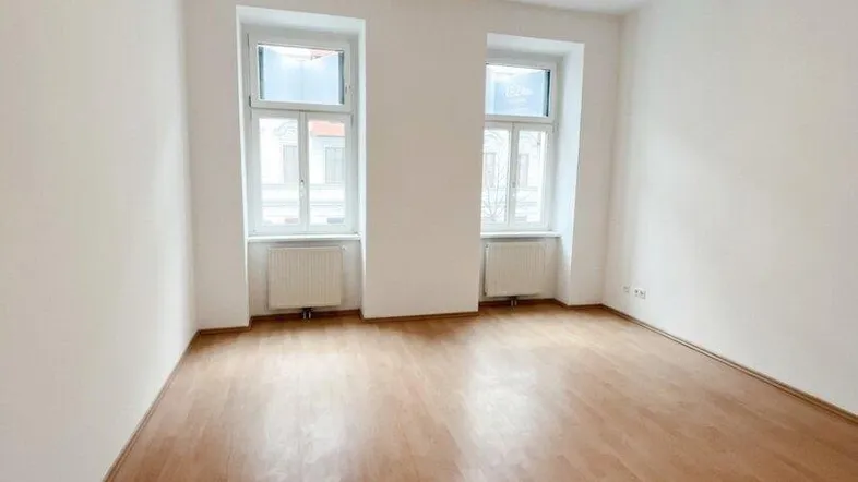 Helles, leeres Zimmer mit Parkettboden und zwei Fenstern, bereit zur individuellen Gestaltung.