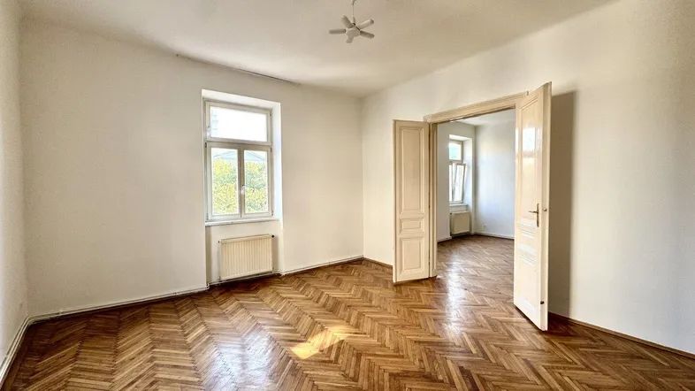 Helles Zimmer mit Fischgrätparkett, hohen Decken und Flügeltüren zu einem weiteren Raum.