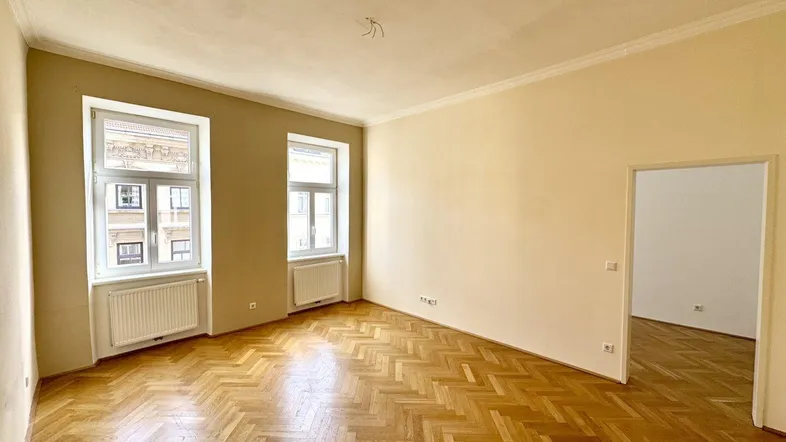 Helles, leeres Zimmer mit Fischgrätparkett, hohen Decken und zwei Fenstern.