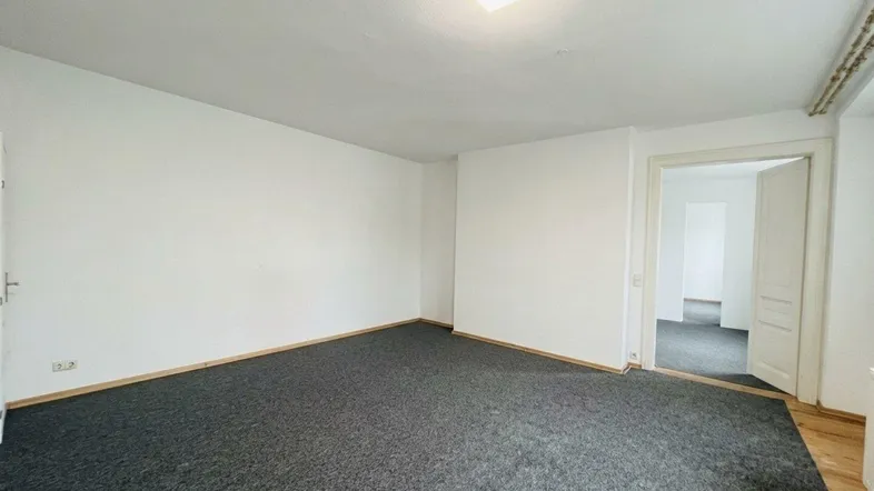 Helles, leeres Zimmer mit dunklem Teppichboden und weißen Wänden, bereit zur individuellen Gestaltung.