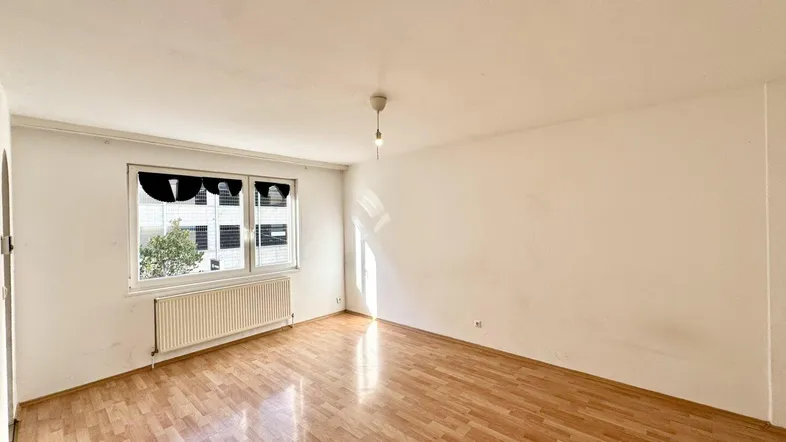 Helles, leeres Wohnzimmer mit Parkettboden und einem Fenster, das Tageslicht hereinlässt.