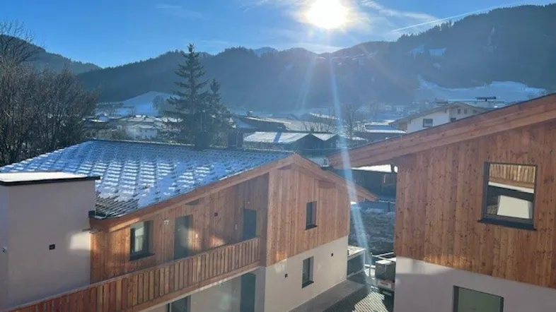 Sonnige Ansicht von modernen Gebäuden mit Holzfassaden und schneebedeckten Dächern vor einer Bergkulisse.