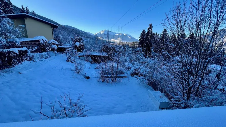 Winterliche Landschaft mit schneebedecktem Garten, Haus und majestätischem Bergblick unter klarem blauem Himmel.