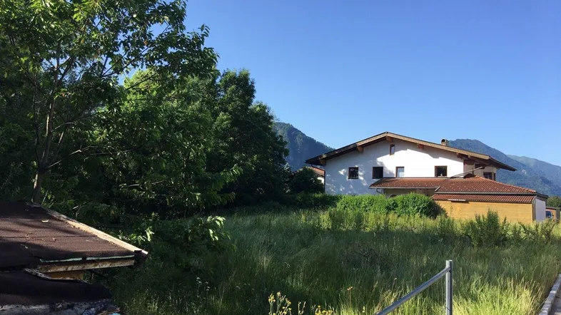 Blick auf ein unbebautes Grundstück mit hohem Gras, umgeben von Bäumen und einem Haus im Hintergrund, mit Bergsicht.