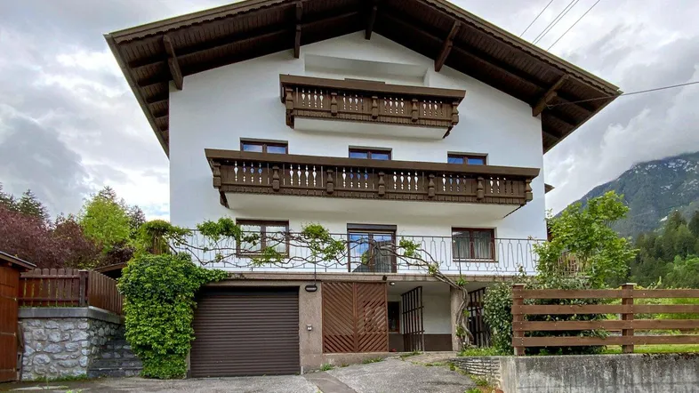 Traditionelles Haus mit weißen Fassaden, Holzbalkonen und integrierter Garage vor bergiger Kulisse.