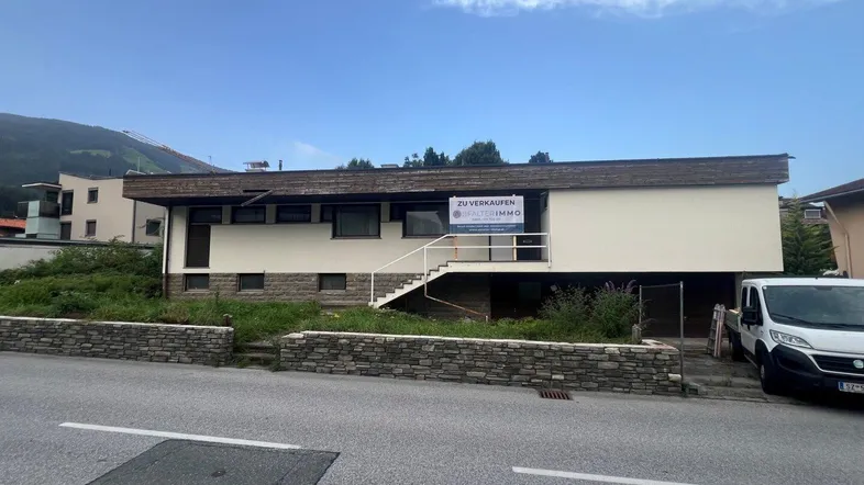 Gebäudeansicht mit einem 'Zu verkaufen'-Schild, umgeben von Grünflächen und mit Bergblick im Hintergrund.