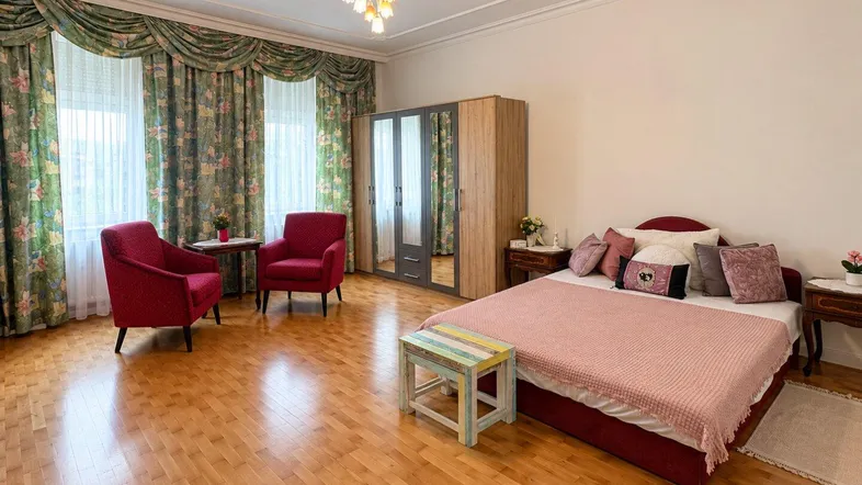 Geräumiges Schlafzimmer mit Holzboden, floralen Vorhängen und zwei roten Sesseln.