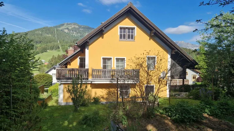 Gelbes Haus mit Satteldach und Balkon, umgeben von grüner Natur und Bergen im Hintergrund.