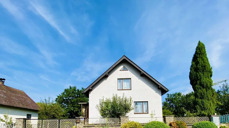 Weißes Einfamilienhaus mit Satteldach, umgeben von einem gepflegten Vorgarten und blauem Himmel.