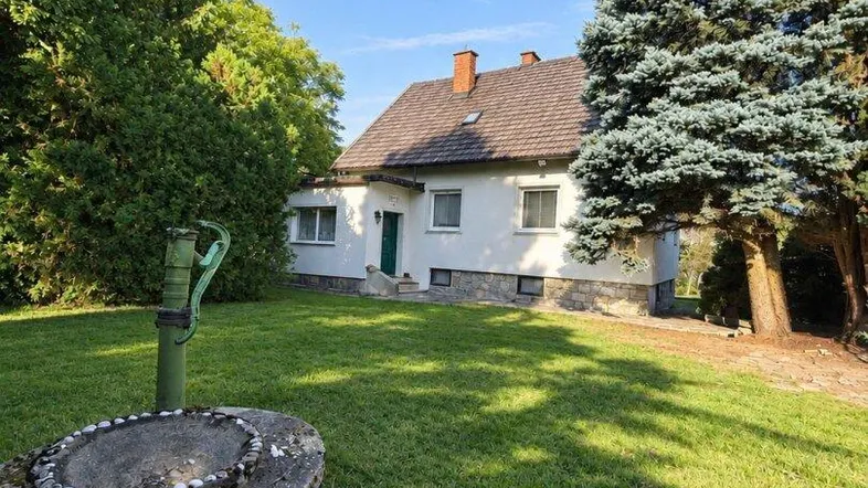 Weißes Einfamilienhaus mit großem grünen Garten und altem Baumbestand unter blauem Himmel.
