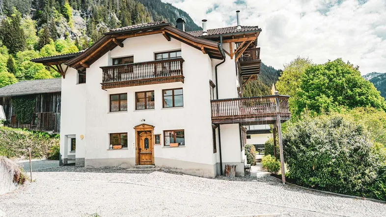 Traditionelles Alpenhaus mit weißer Fassade und Holzbalkonen, umgeben von grünen Bergen.