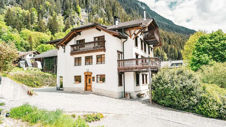 Traditionelles Alpenhaus mit Holzbalkonen und Blick auf die bewaldeten Berge unter bewölktem Himmel.