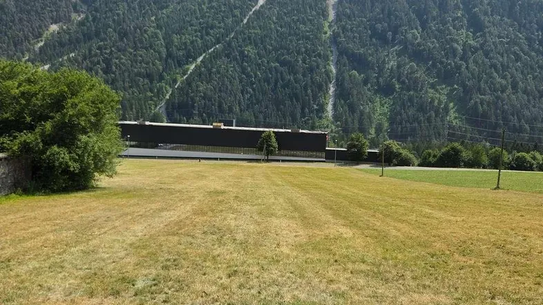Weitläufiges Feld mit Bergblick und einem Gebäude im Hintergrund unter bewölktem Himmel.