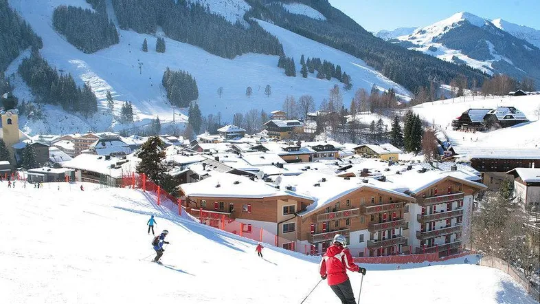 Panoramablick auf ein verschneites Skigebiet mit Gebäuden und Skifahrern unter blauem Himmel.