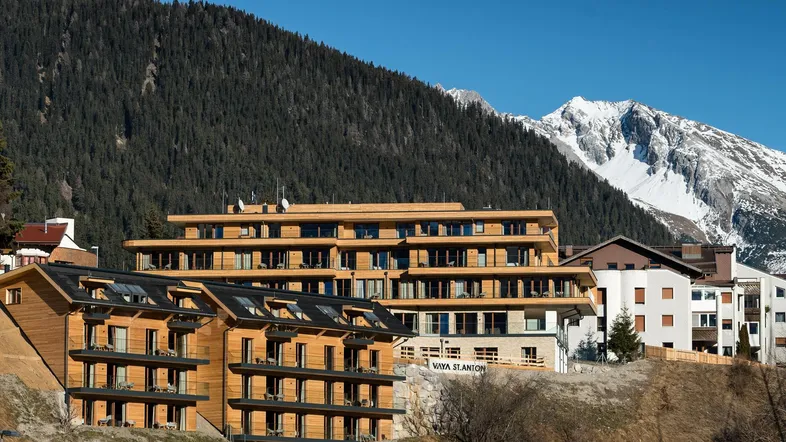 Moderne Holzfassaden von Gebäuden in einer alpinen Landschaft mit schneebedeckten Bergen im Hintergrund.