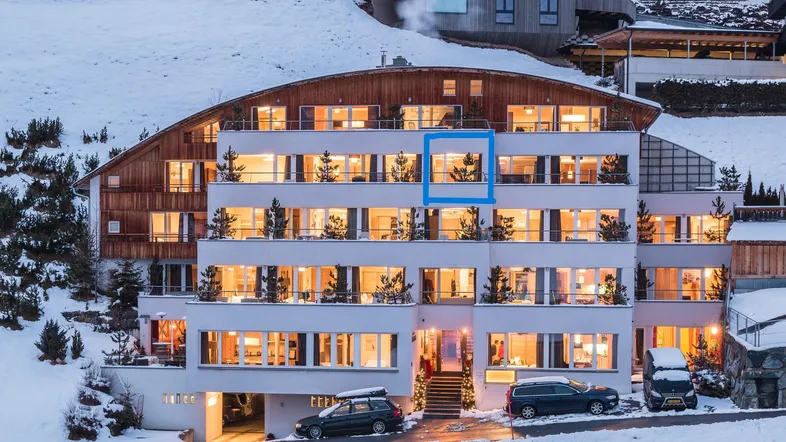 Modernes Apartmentgebäude in den Bergen, beleuchtet bei Nacht mit Schnee und vielen Balkonen.