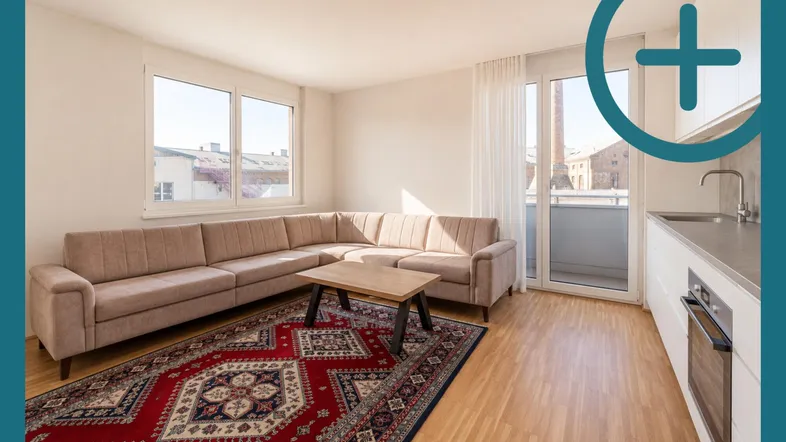 Helles Wohnzimmer mit beigem Sofa, Holzboden und integrierter Küchenzeile mit Zugang zum Balkon.