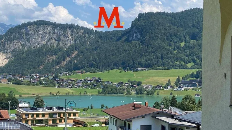 Panoramablick auf den See und die umliegenden Berge unter blauem Himmel mit leichter Bewölkung.