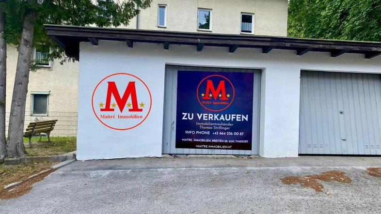 Weiße Garage mit zwei Toren und einem 'Zu verkaufen'-Schild, davor eine asphaltierte Fläche und im Hintergrund ein Wohngebäude.