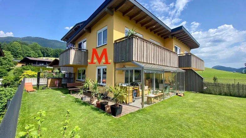 Gelbes Wohnhaus mit Holzbalkonen, Wintergarten und gepflegtem Garten in bergiger Umgebung.