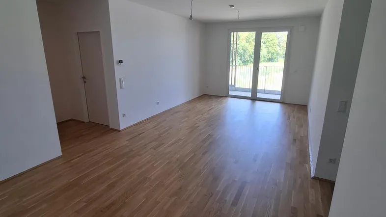 Helles, leeres Wohnzimmer mit neuem Parkettboden und Zugang zu einem Balkon.