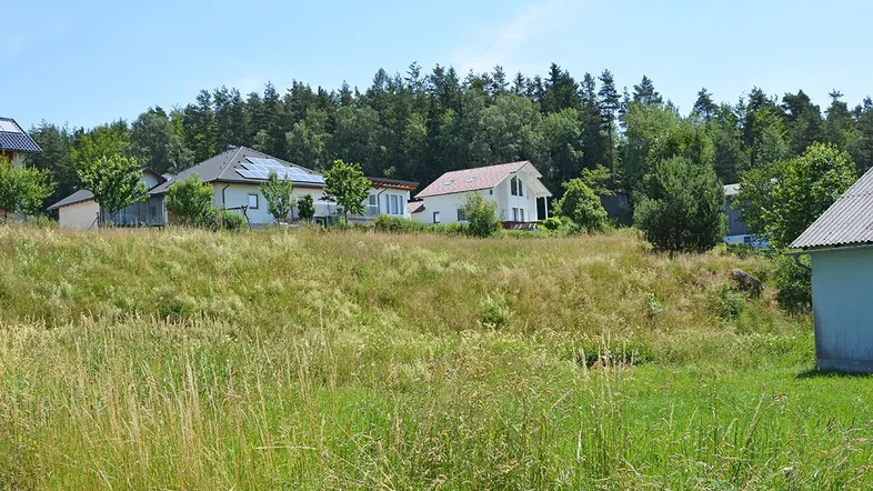 Ländliche Umgebung mit Häusern auf einem Hügel und Wald im Hintergrund unter blauem Himmel.