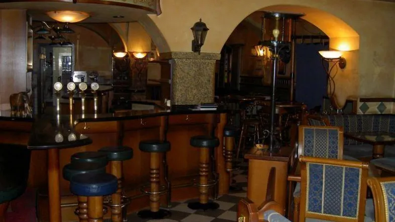 Traditionelle Bar mit Holztresen, Barhockern und gemütlichen Sitzbereichen unter Gewölbebögen und Fliesenboden.