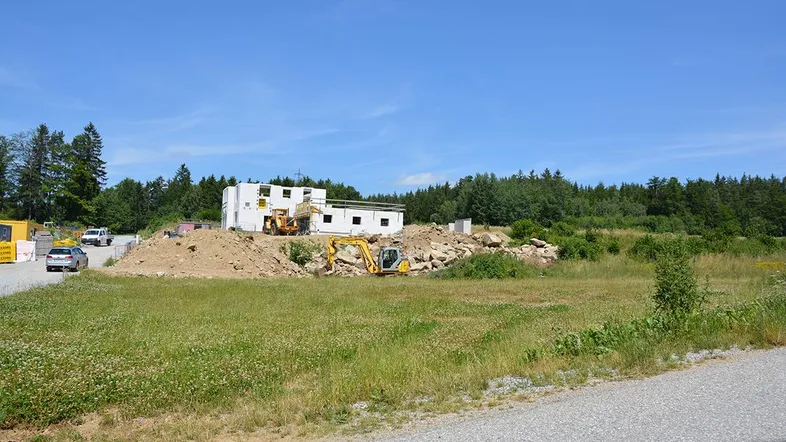 Baustelle mit im Bau befindlichem Gebäude, umgeben von grünen Feldern und Wald unter blauem Himmel.