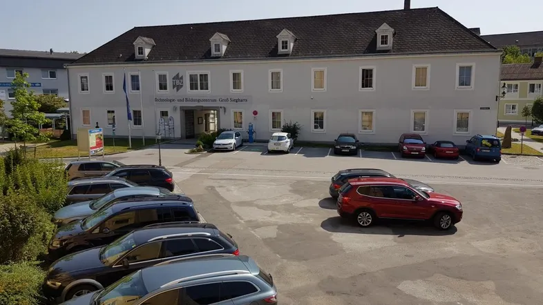 Großes Gebäude mit mehreren Fenstern und einem Parkplatz davor unter blauem Himmel.