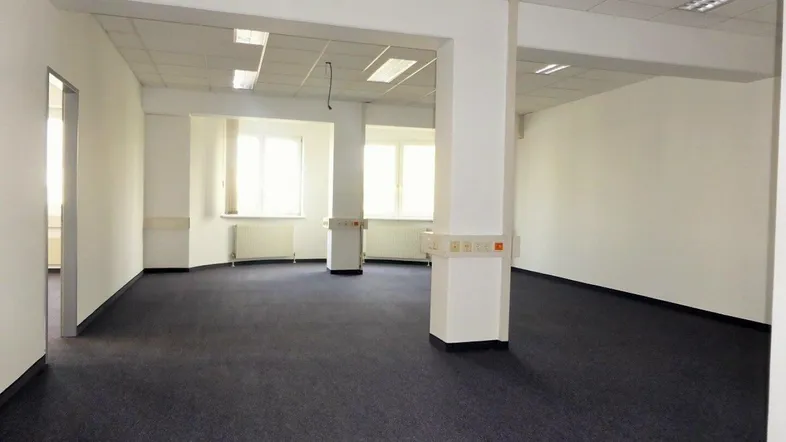 Geräumiges Büro mit Teppichboden, weißen Wänden und Deckenplatten, sowie großen Fenstern für viel Tageslicht.