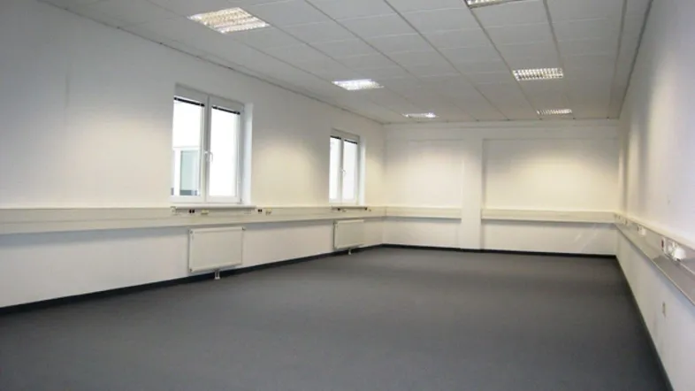 Geräumiges, leeres Büro mit weißen Wänden, grauen Teppichböden und großen Fenstern, bereit zur Einrichtung.
