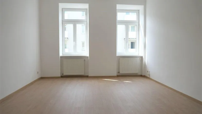 Helles, leeres Zimmer mit zwei Fenstern, Heizkörpern und neuem Laminatboden, bereit zur Einrichtung.