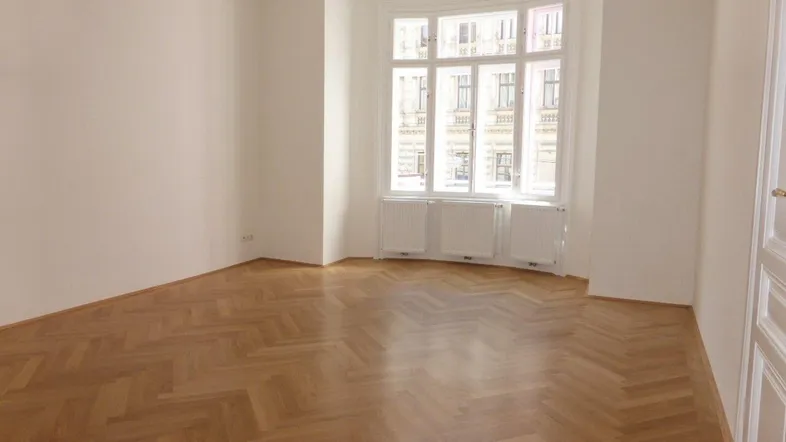 Helles Zimmer mit Fischgrätparkett und einem großen Erkerfenster, das viel Tageslicht hereinlässt.