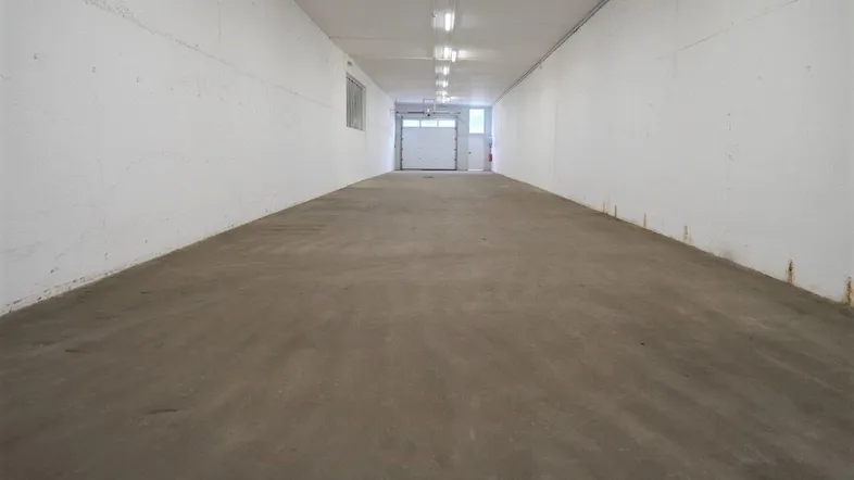 Langer, leerer Lagerraum mit Betonboden, weißen Wänden und Reihen von Deckenleuchten.