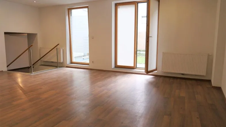 Heller, leerer Raum mit Holzboden, großen Fenstern und Zugang zu einem Außenbereich.