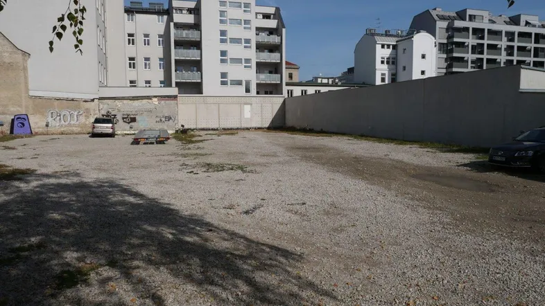 Große, unbefestigte Fläche, die als Parkplatz dient, umgeben von städtischen Gebäuden unter blauem Himmel.