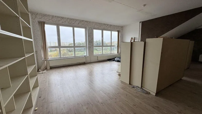 Geräumiger Innenraum mit großen Fenstern, Laminatboden und eingebauten Regalen, der Renovierungsbedarf aufweist.